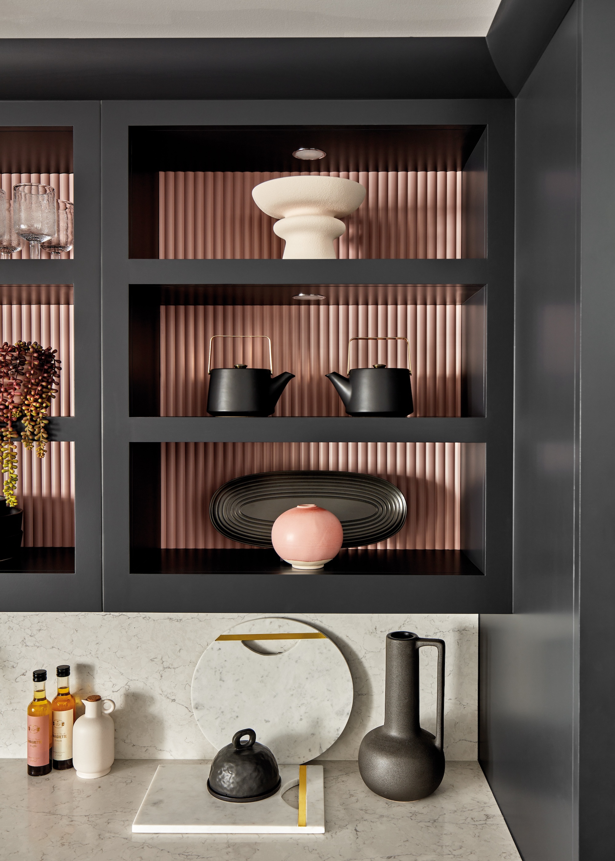 A dark open shelved kitchen with decor pieces inside as well as a light colored countertop with a water vessel, chopping board and other kitchen essentials.
