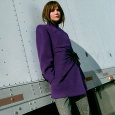Promo image for Emilia Jones' Ones to Watch story. In the image, she is leaning against a white semi-truck wearing a purple coat and gray trousers. 