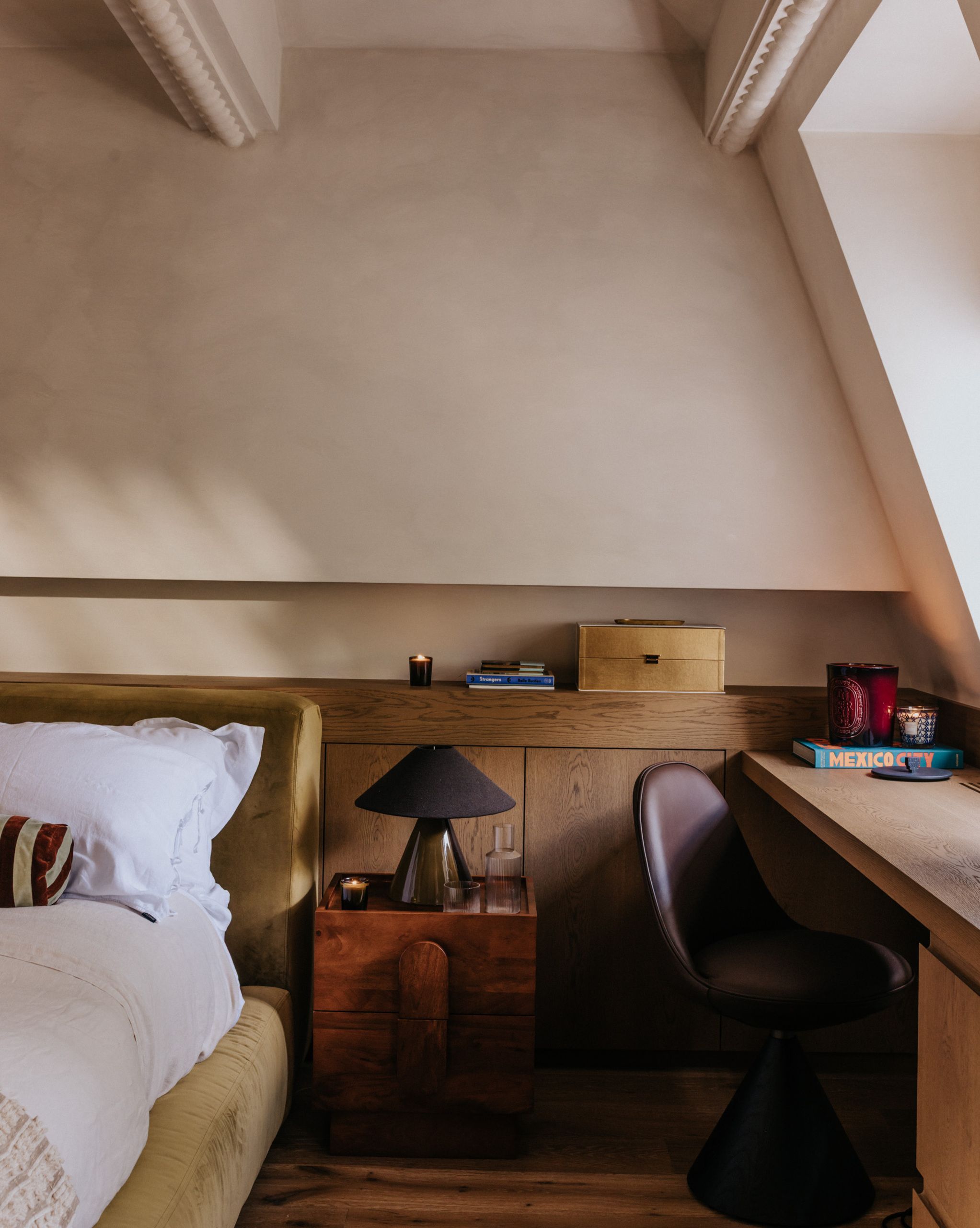 Image of a limewash beige bedroom with wooden built-in cabinetry, an olive bed frame, white bedding, and a desk with a purple leather chair.