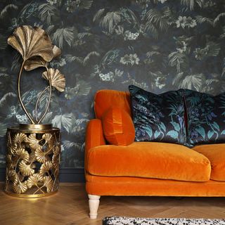 A living room with a dark floral-print wallpaper and an orange velvet sofa