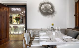 Dining area in the restaurant showcasing leather cream chairs on the left and grey sofa on the right with a long table dressed with napkins, wine glasses and cutleries and a yellow rose in the middle. White wall with skull drawn art
