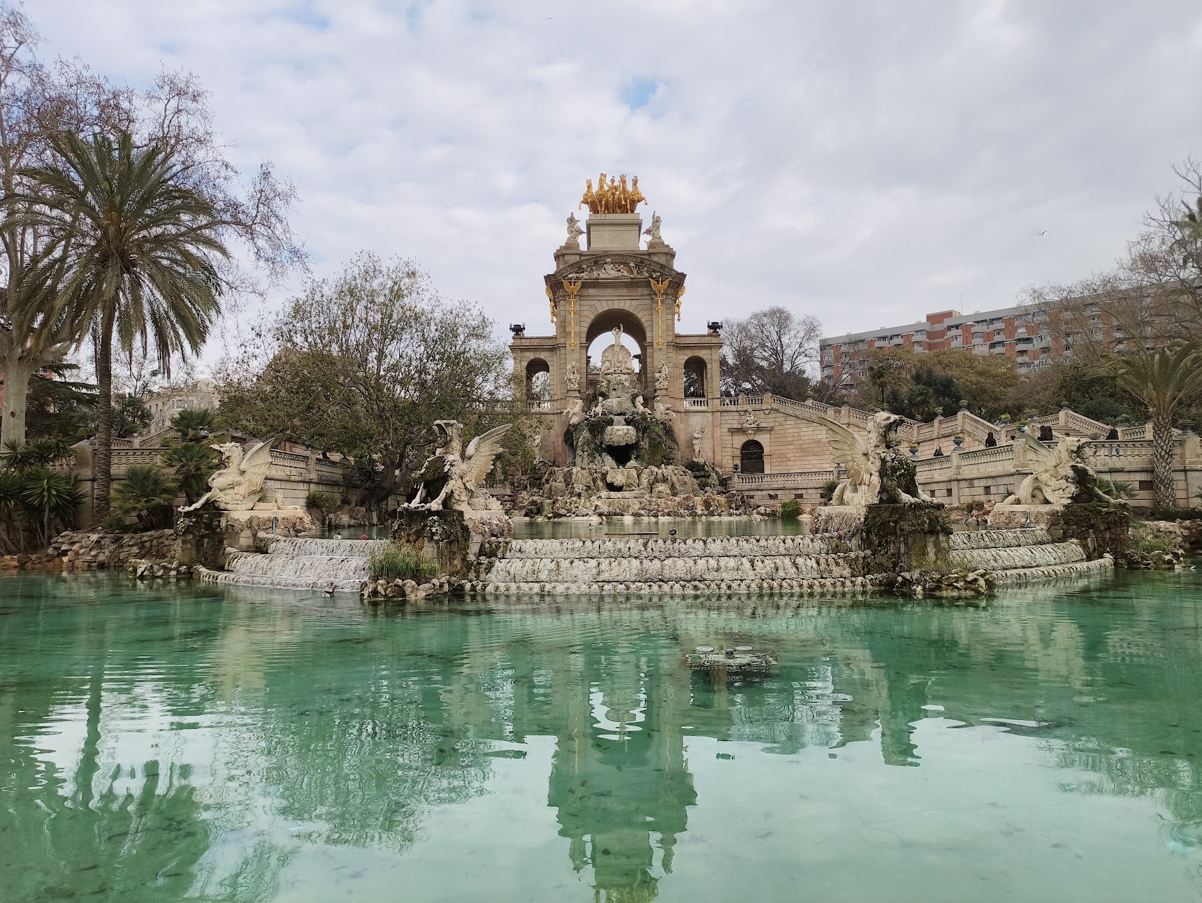 A grand, multi-tiered stone fountain features a golden chariot sculpture at its peak and winged dragon statues flanking a turquoise pool of water.