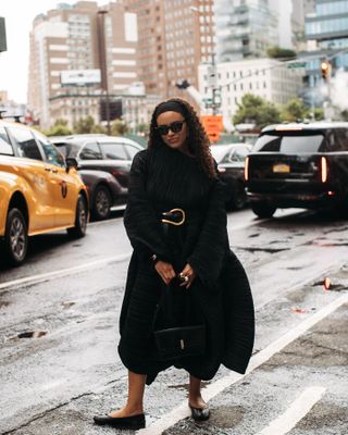 Woman wearing black pleated set, black handbag, and black Tabi ballet flats while standing on New York City street.