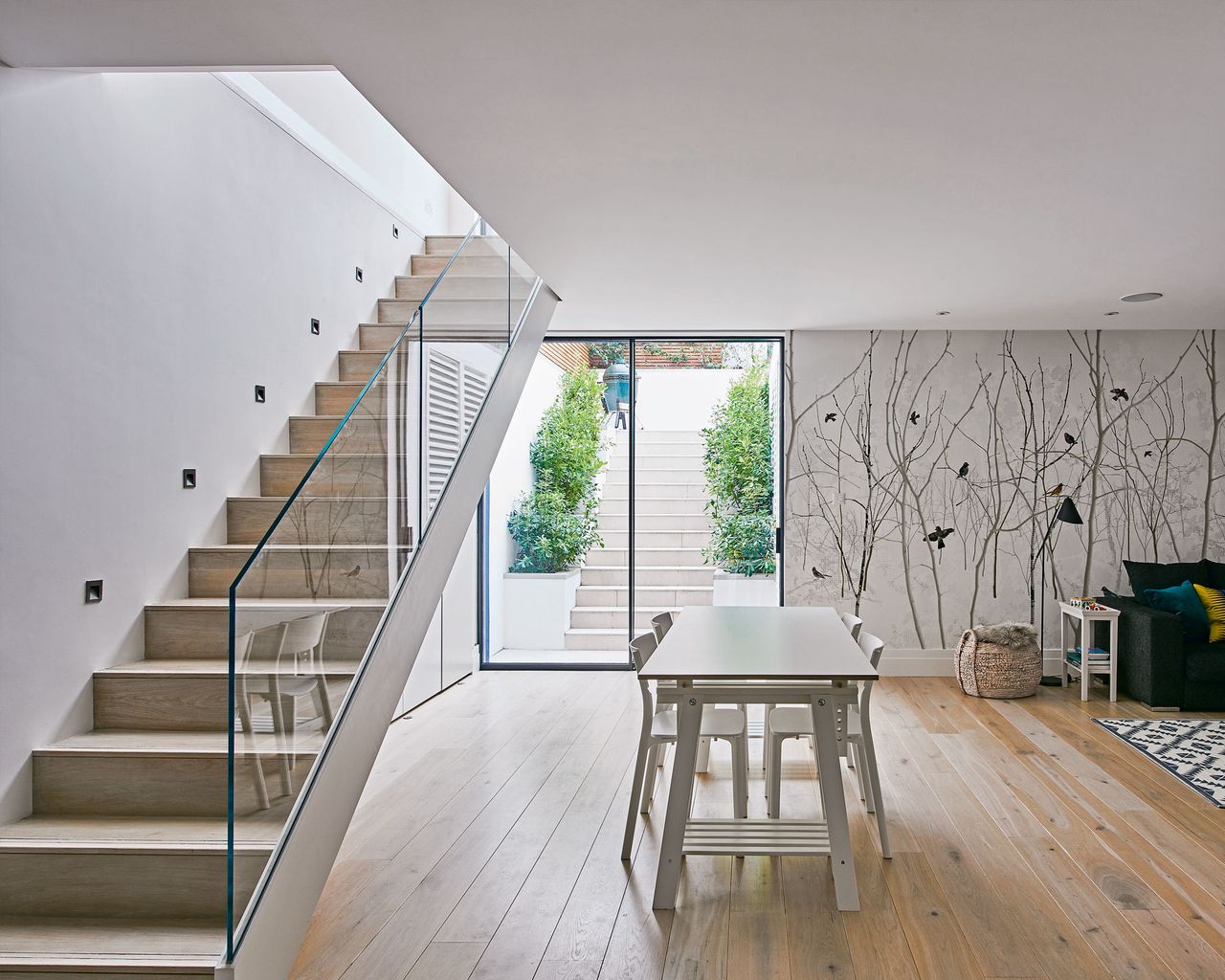 Basement room with staircase with sheet glass bannister, glass door to the garden, wooden flooring and large dining table with chairs.