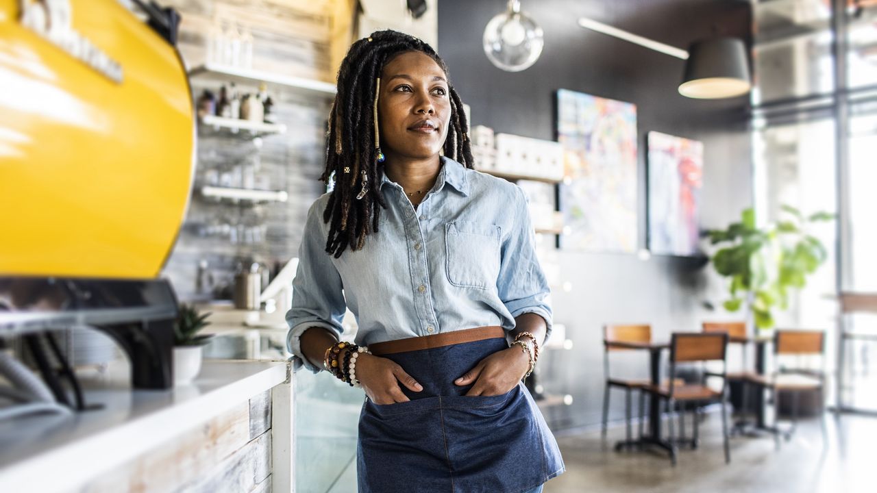 A small business owner stands at the counter of a café, looking thoughtful.