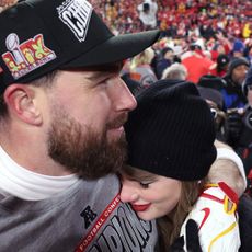 Travis Kelce hugging Taylor Swift, wearing a black knit hat, on the field after the AFC Championship game