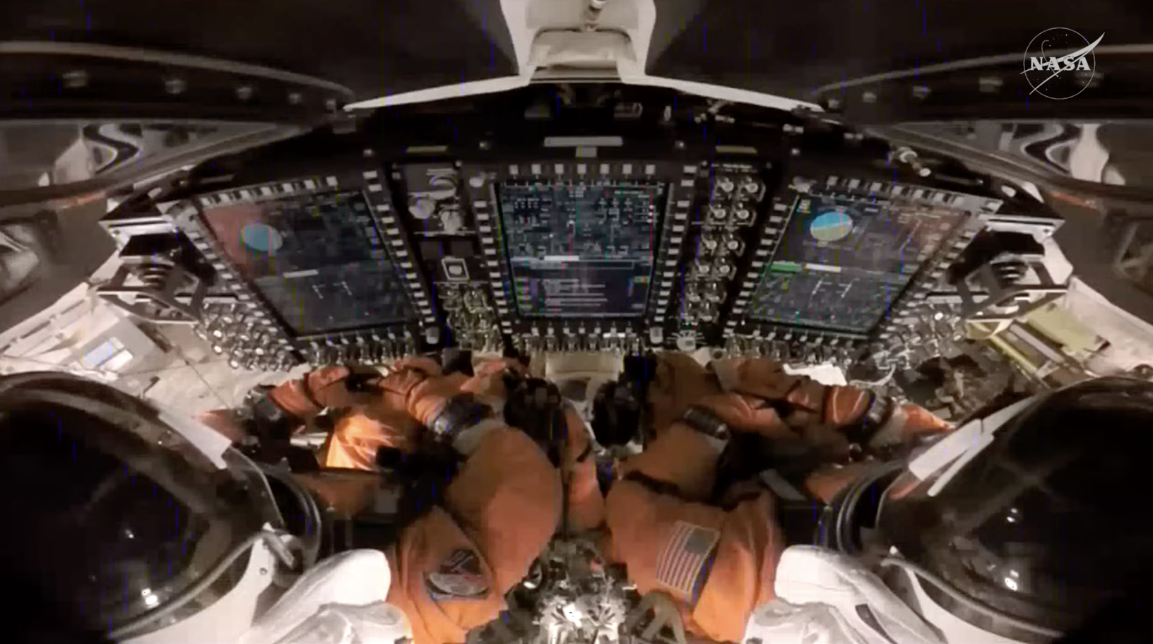 Two people sitting in orange suits look at large screen monitors