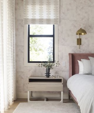 Nightstand with neutral rug in front and bed alongside in bedroom with neutral patterned wallpaper and brass wall light