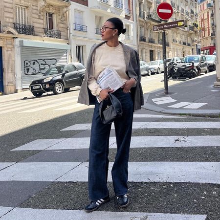 French fashion influencer Léna Farouil poses on a Paris sidewalk wearing eyeglasses, chunky earrings a beige sweatshirt, houndstooth blazer, dark-wash jeans, a black mini bag, and black Adidas Samba sneakers