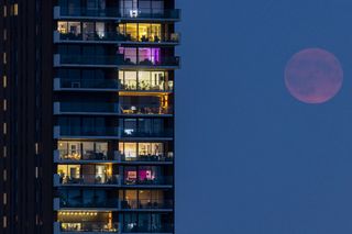 a hazy red moon faintly shines to the right of a tall apartment block building.