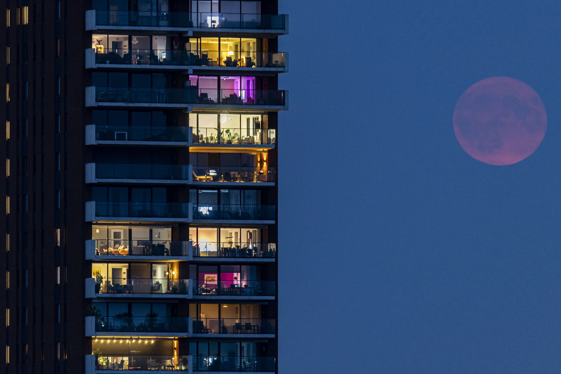 To the right of a tall apartment block, a hazy red moon glows faintly.