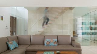 Modern living room with a minimalist floating staircase behind a glass wall, featuring a sleek leather sectional and decorative cushions. 