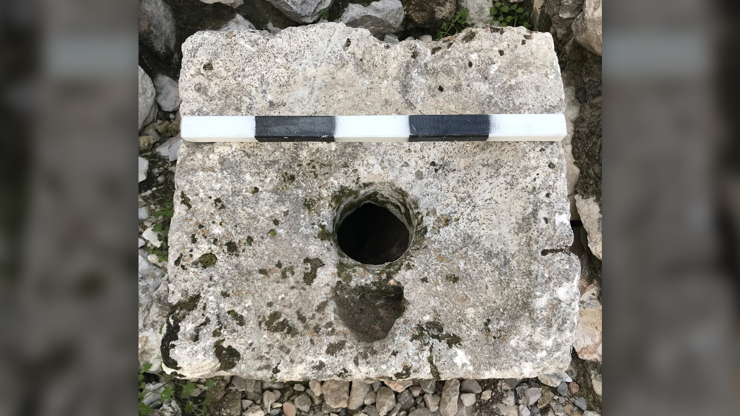 Wir sehen einen weiß-schwarzen Steinblock mit einem Loch in der Mitte.  Daran ist ein Messstab angebracht.