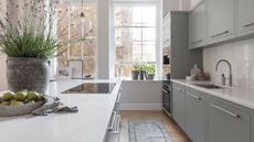 A naturally lit kitchen with very large windows alone the back wall, with light blue cabinets and and island. White countertops. 