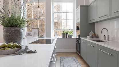 A naturally lit kitchen with very large windows alone the back wall, with light blue cabinets and and island. White countertops. 