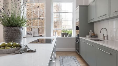 A naturally lit kitchen with very large windows alone the back wall, with light blue cabinets and and island. White countertops. 
