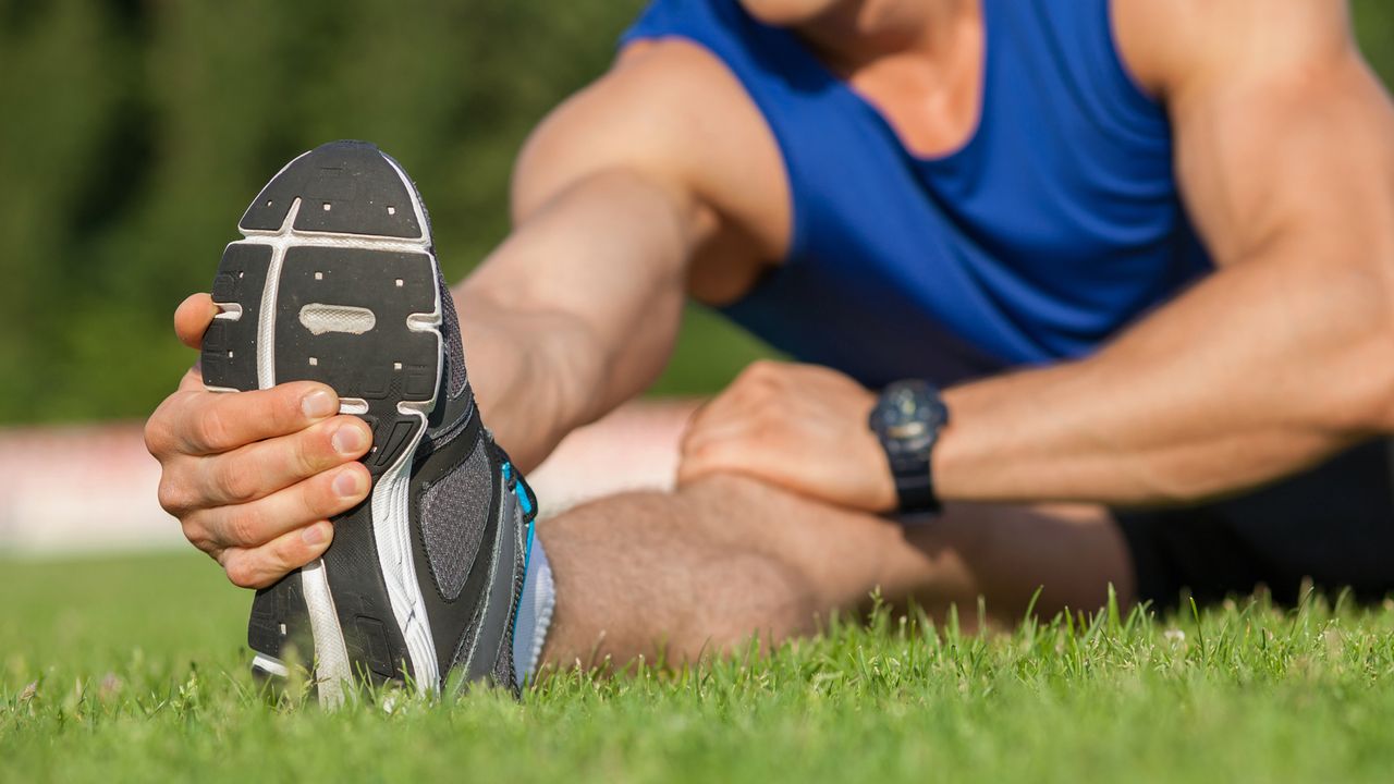 Close-up of leg of male runner stretching his leg forward. He is touching it with his hand and warming up