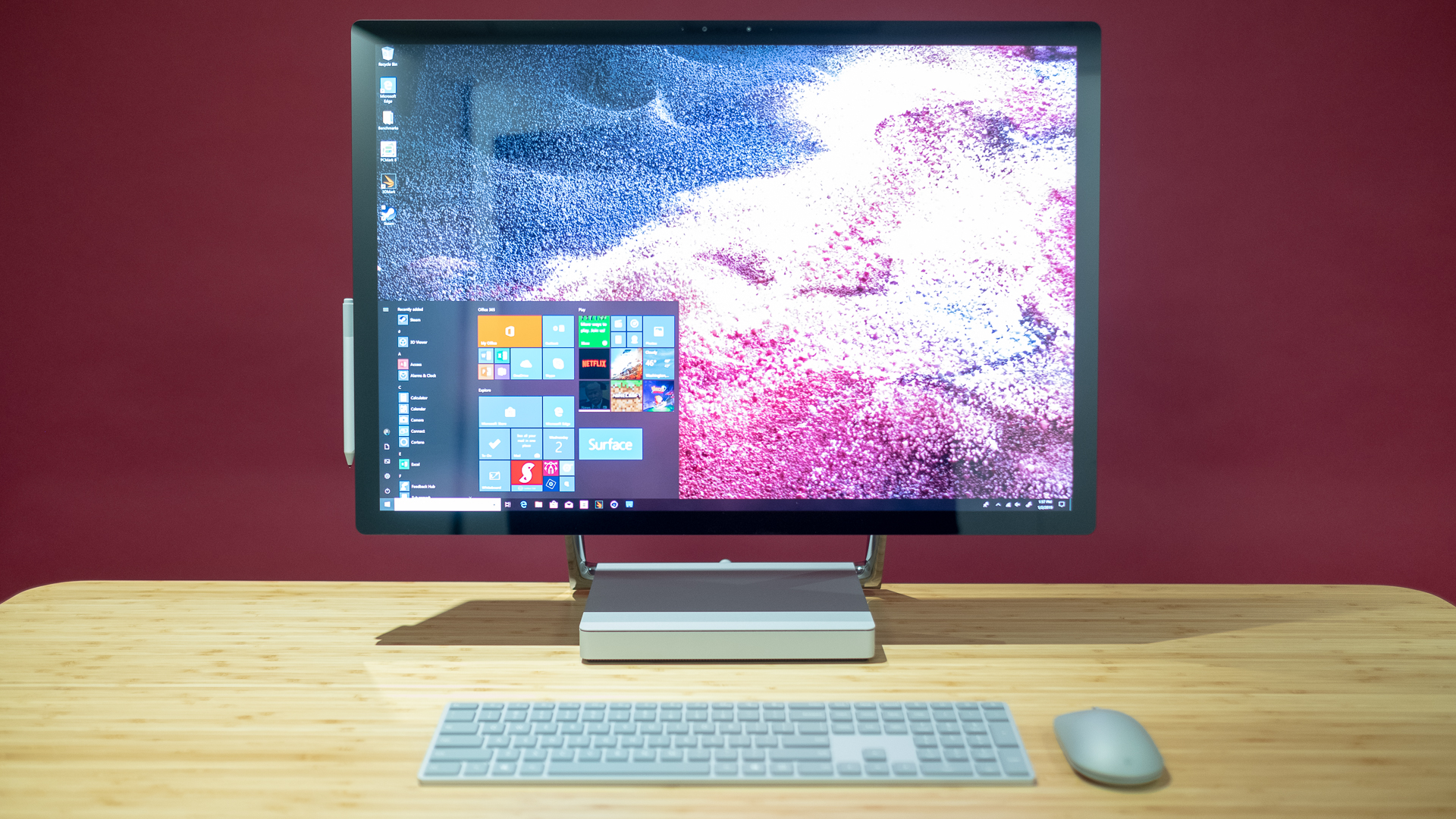 Surface Studio 2 on a table with its keyboard and mouse against a maroon wall