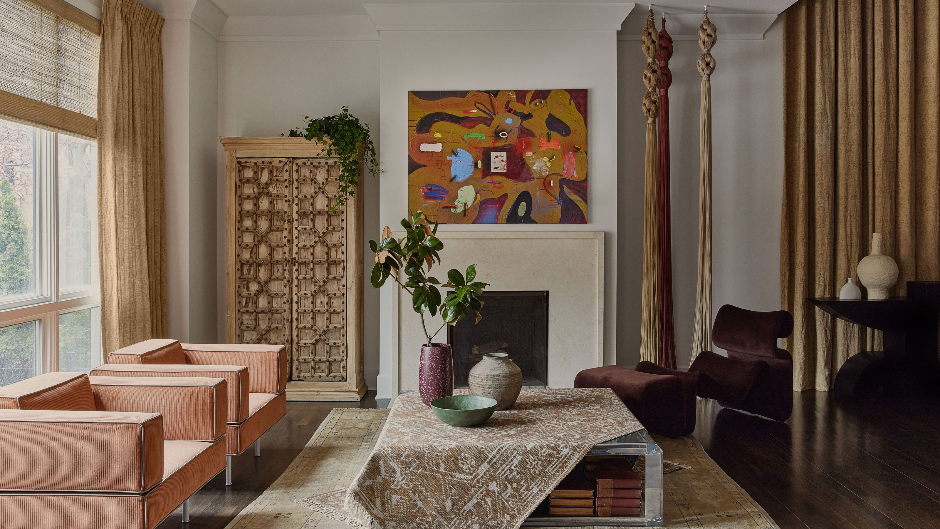 A living room with salmon armchairs, a plum-toned accent chair, two long tassels attached to the ceiling, a carpet strewn on the coffee table and a vintage artwork 