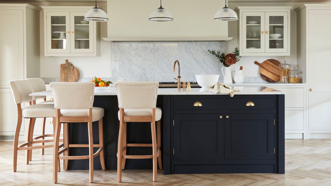 A kitchen with a marble island with a black base, a bowl of tomatoes on it and four cream and wood bar stools around the left corner of it, two glass pendant lights on the ceiling, and cream cabinetry