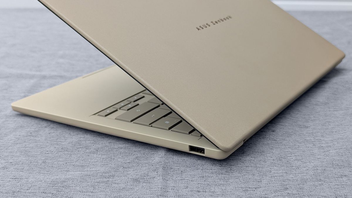 Partially open beige ASUS Zenbook A14 on gray fabric, showing keyboard and a single USB port on the right side.