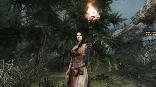 A woman holds a torch while standing in the woods in Skyrim.