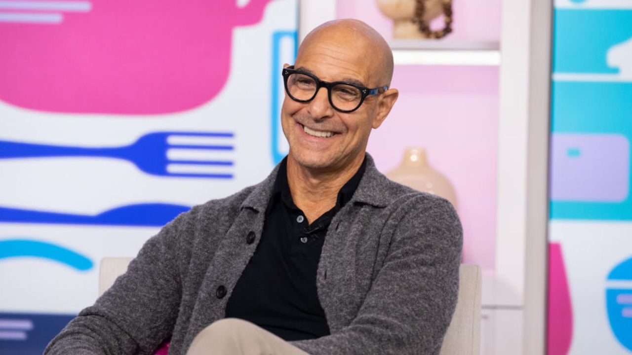 Stanley Tucci wearing a grey jumper, sitting on a chair with a colourful background