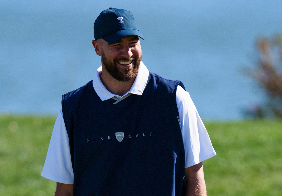 Travis Kelce wearing a navy cap, navy vest and white polo on a golf course