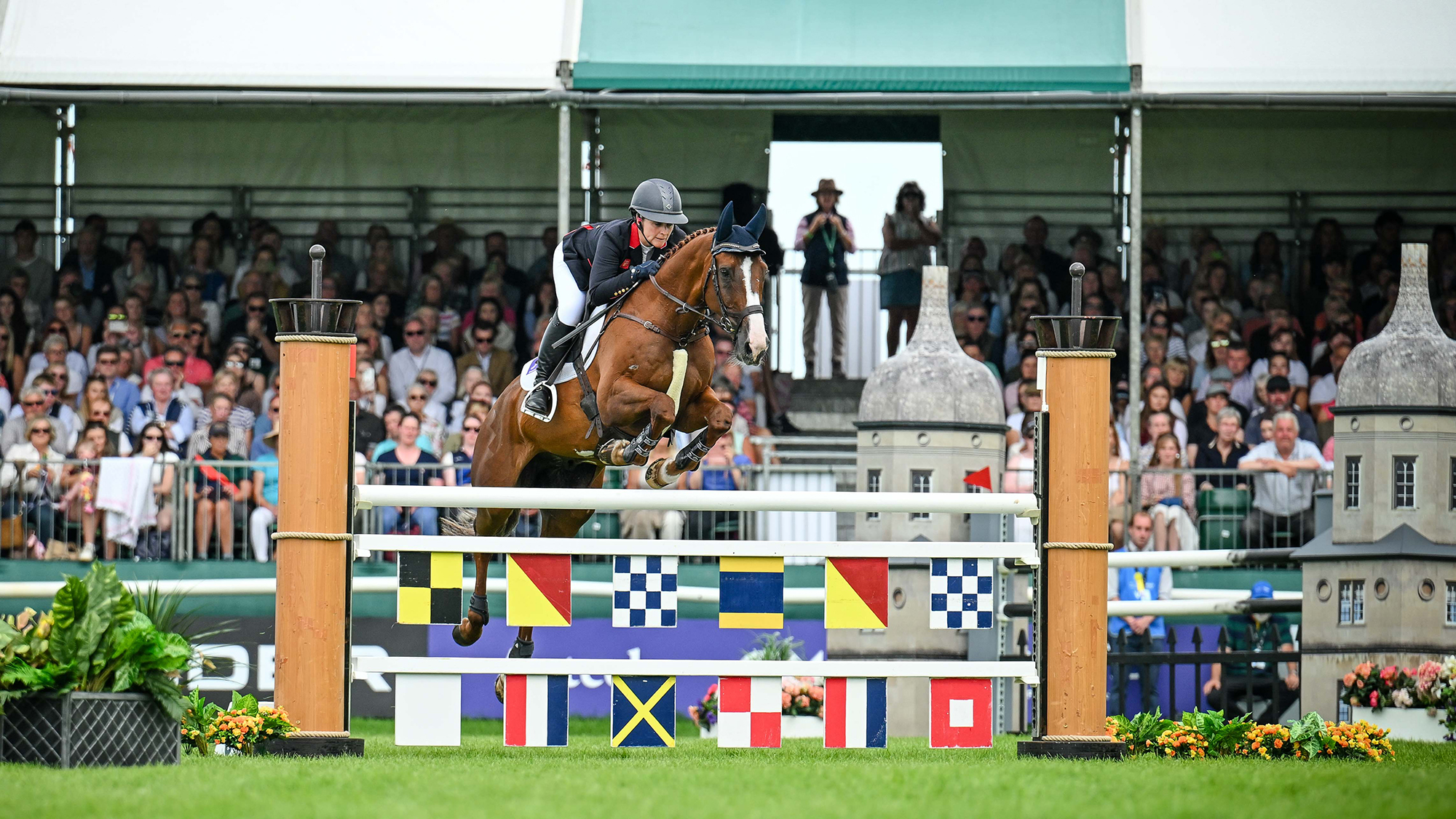 Gemma Stevens, a rider in a black coat, rides chestnut horse Chilli Knight over a fence decorated with flags at Burghley 2025