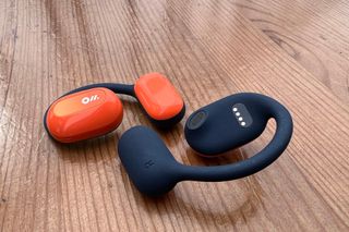 The right Oladance open ear headphone has been turned over to see the blue underside next to the orange left headphone on a wooden surface