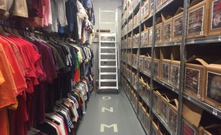storage of 3 clothing rails on the left and boxes on shelving on the right with a ladder leaning on the wall at the end