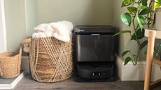 A black Eureka J15 Pro Ultra robot vacuum next to a woven basket and a leafy green plant, with a sage green wall behind it
