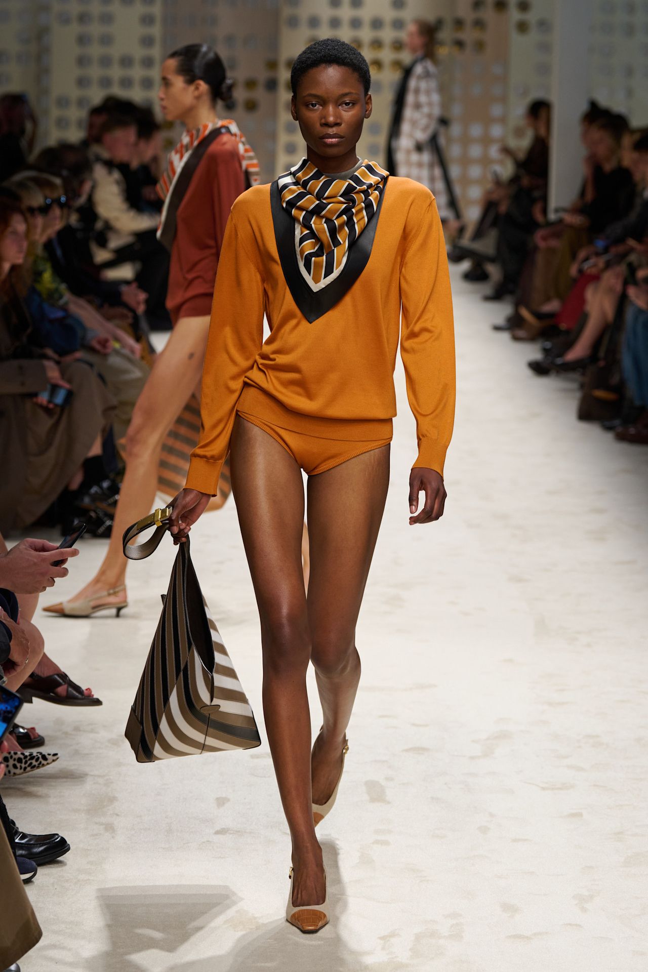 A Tod's model wearing a tangerine-hued crewneck sweater styled with matching briefs, two-tone pointed pumps, a striped leather tote bag, and a black, white, and orange silk scarf around the neck.