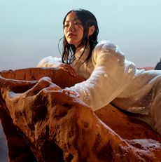 Anna Sawai reclines on red desert rocks in a dark textured top and a shimmering skirt.