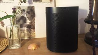 The Sonos One speaker in black pictured on a shelf next to a vase and a shell