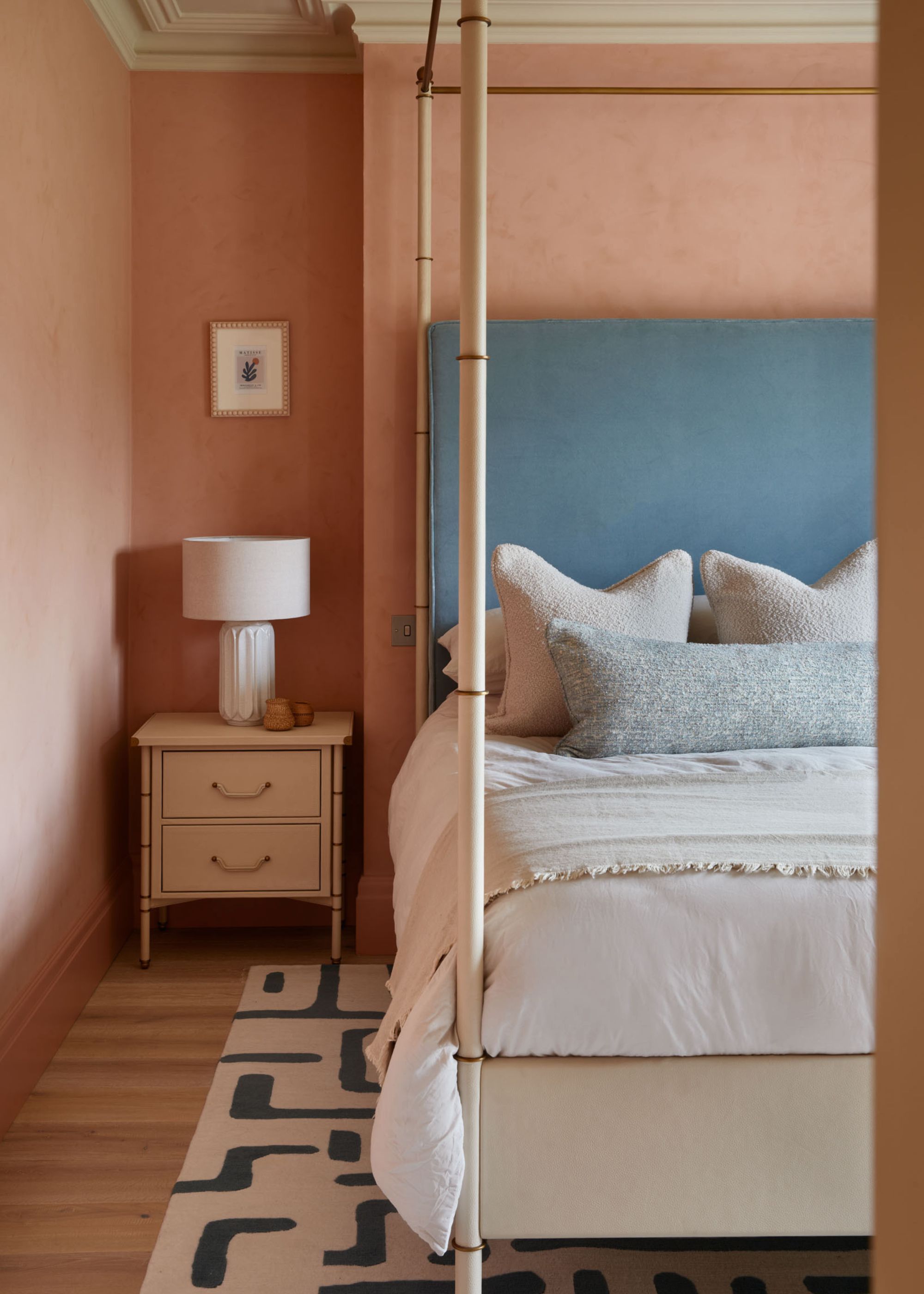 A bedroom with peachy-pink walls, a four-poster bed with a light blue headboard and white, layered bedding, and a pale blue accent cushion. A black and white patterned rug on the floor, and a cream nightstand and lamp.