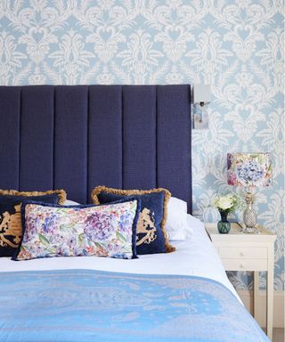 Bedroom wall light ideas showing a bed with a dark blue headboard and blue and white bedding next to a floral bedside lamp and discreet wall light