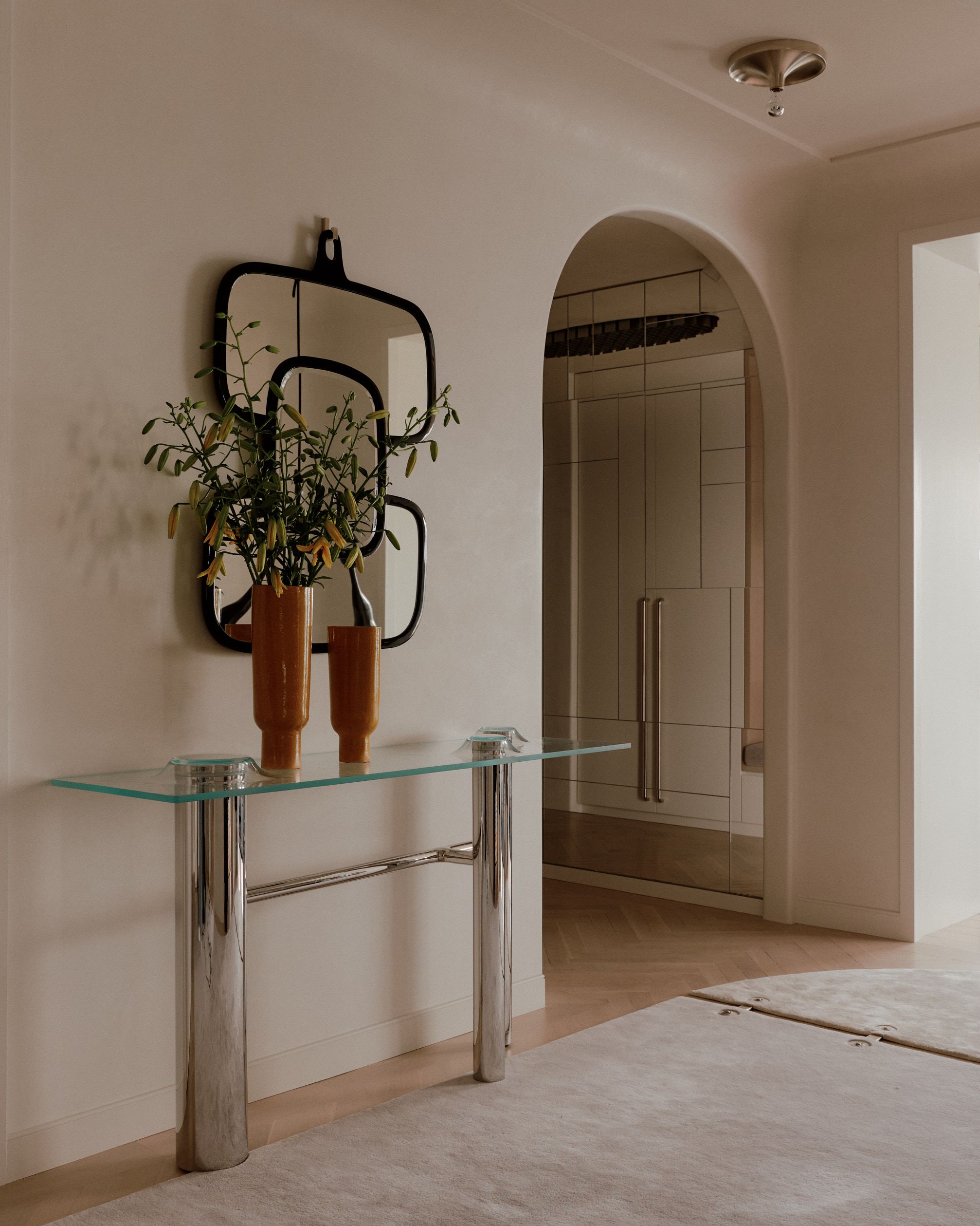 entryway console with metallic and glass table with two vases and a mirror, beige rug, and arched doorway leading through to a hallway with mirrored walls