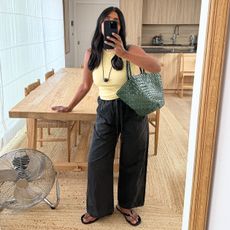 British influencer Monikh Dale poses for a mirror selfie in her London living room with a wood table, rattan chairs, and jute rug while wearing a butter-yellow tank top, woven green tote bag, cord necklace with a black pendant, black linen pants, and flip-flop sandals