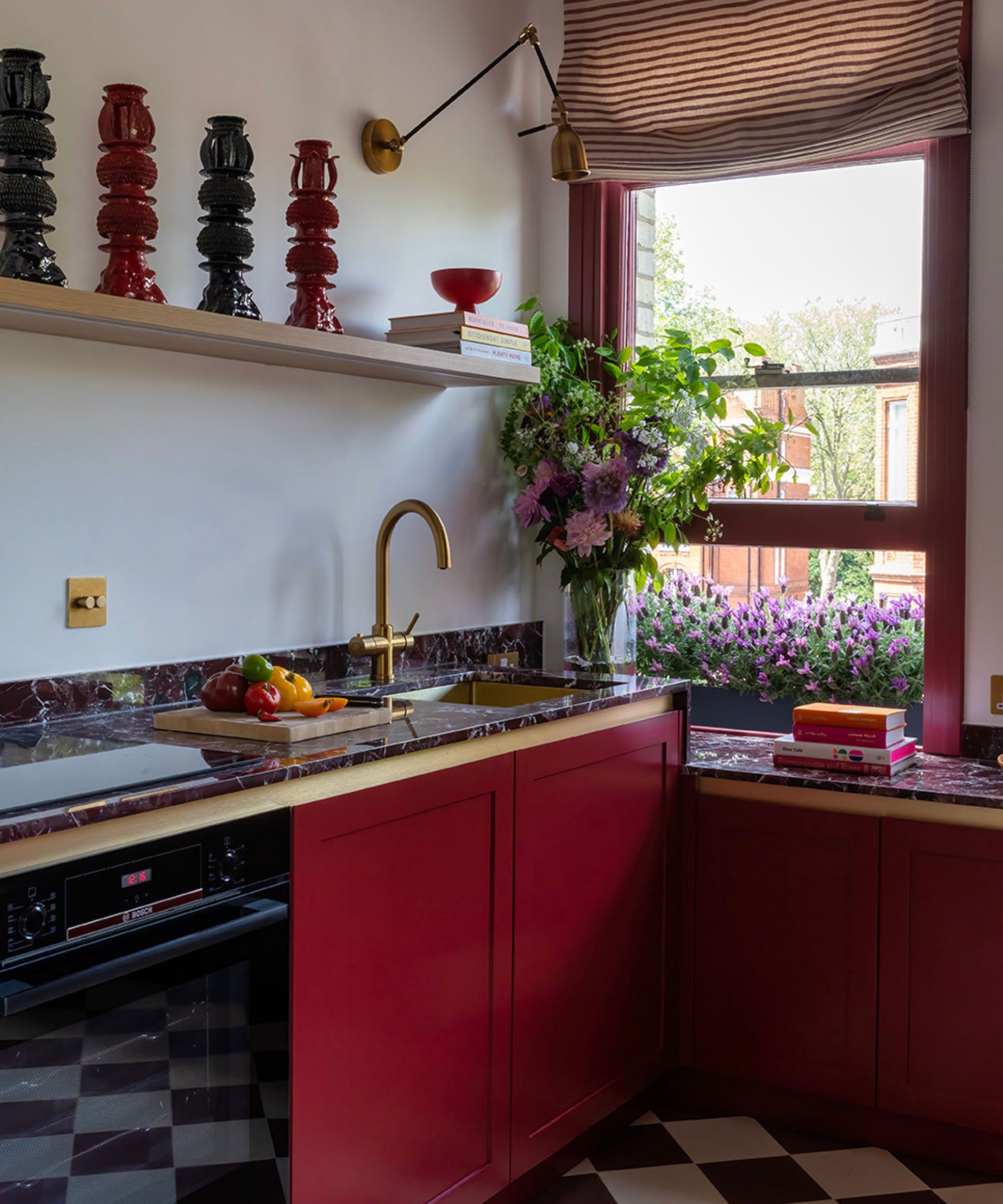 Compact kitchen with bold red cabinets, brass tap and dark marble worktops. A striped Roman blind frames the window, while flowers and colourful accessories brighten the space, creating a vibrant, cosy design with strong colour contrast.