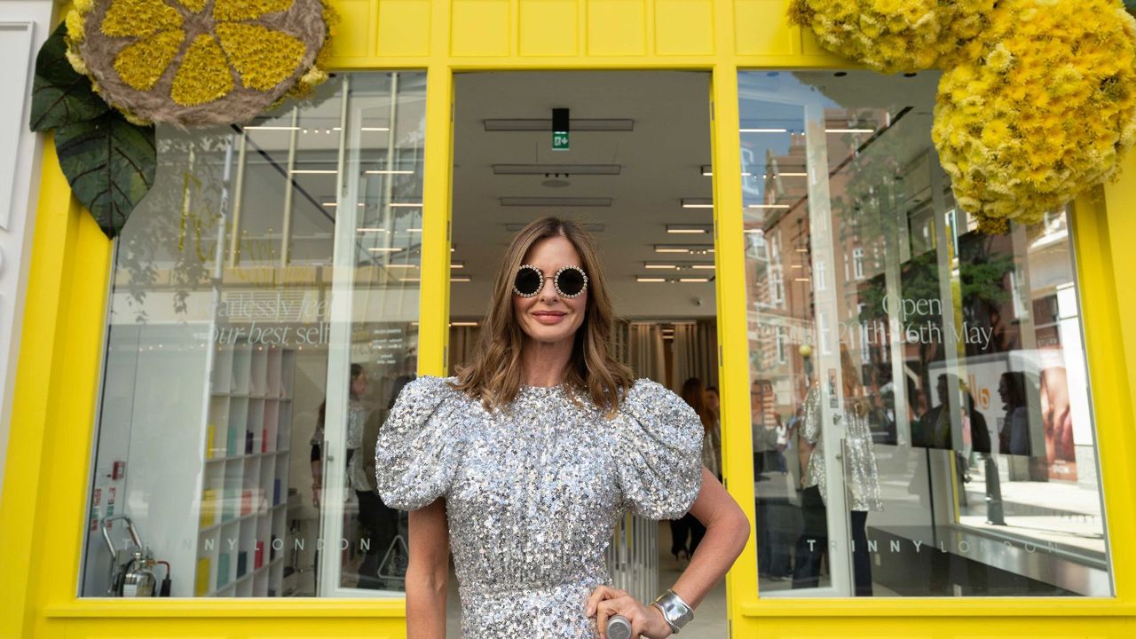 Trinny Woodall standing in front of Trinny London store wearing metallic dress and large round sunglasses