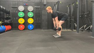 Fitness writer Harry Bullmore performs a Romanian deadlift in a gym. He holds dumbbells at mid-shin height as he hinges forward from his hips, maintaining a straight back. Behind him we can see colorful weight plates and gym equipment.