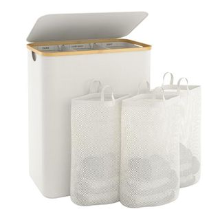 A cream laundry basket with a bamboo trim and flip lid, with three removable mesh bags in front of it. 