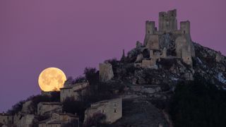 The full moon sets in the lower left of the image behind the village with the Rocca Calascio fortress perched on top of the hill on the right. The sky is a hazy pink/purple in color. 