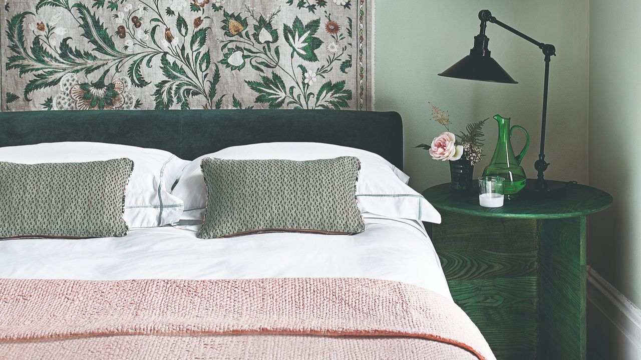 A green-painted bedroom with a bed with a green velvet headboard and a matching bedside table, complemented by a pink throw on the bed