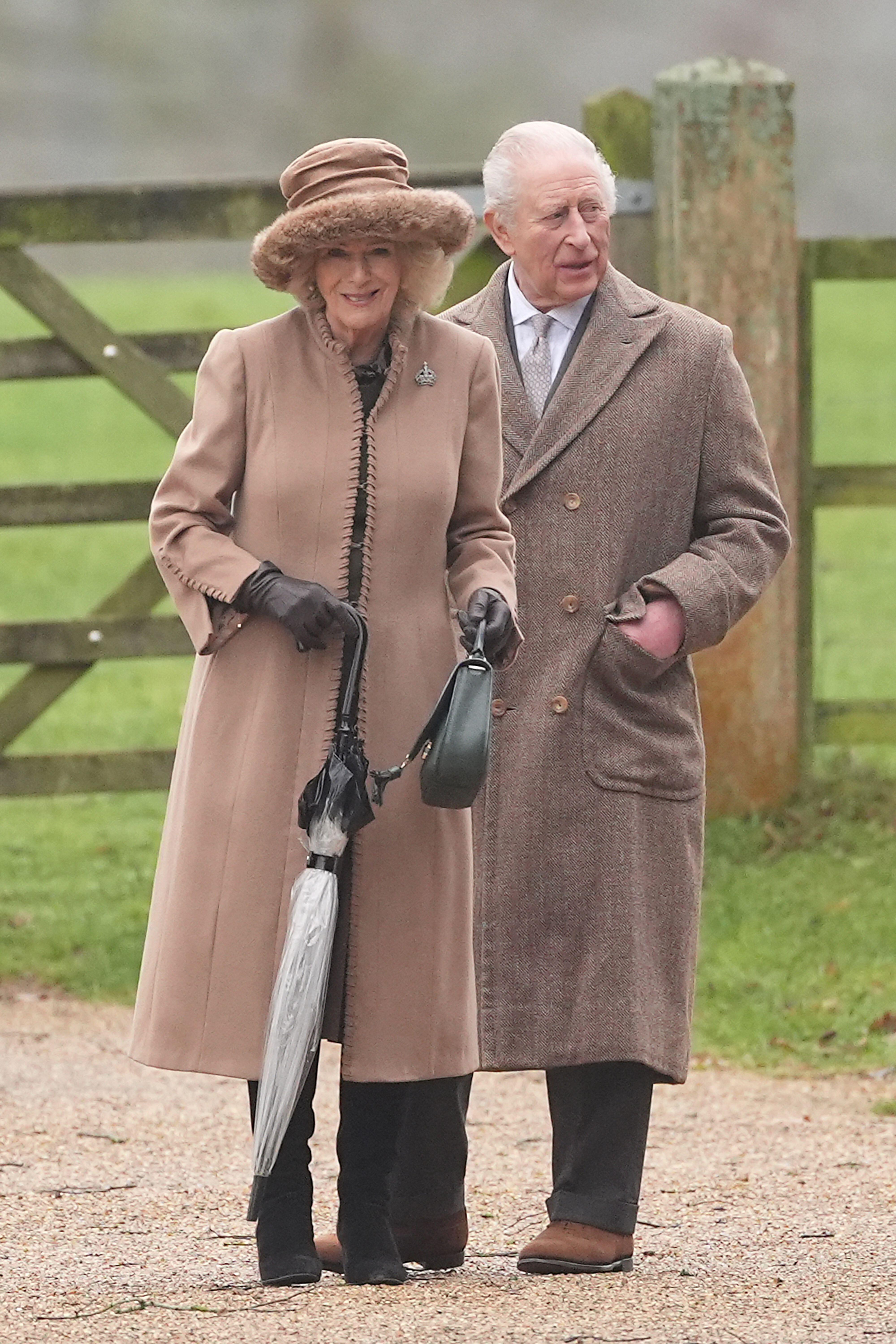 Queen Camilla wearing a tan coat and hat smiling next to King Charles at church