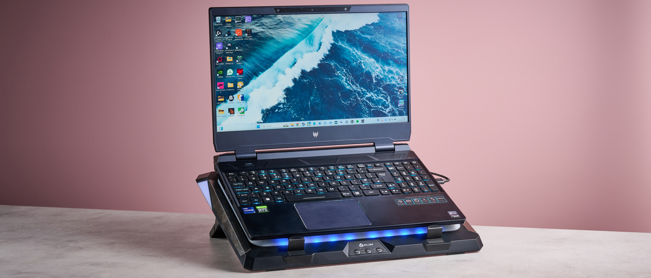 An Acer Predator Helios 300 laptop sat on top of the Klim Mistral laptop cooling pad in front of a pink background.