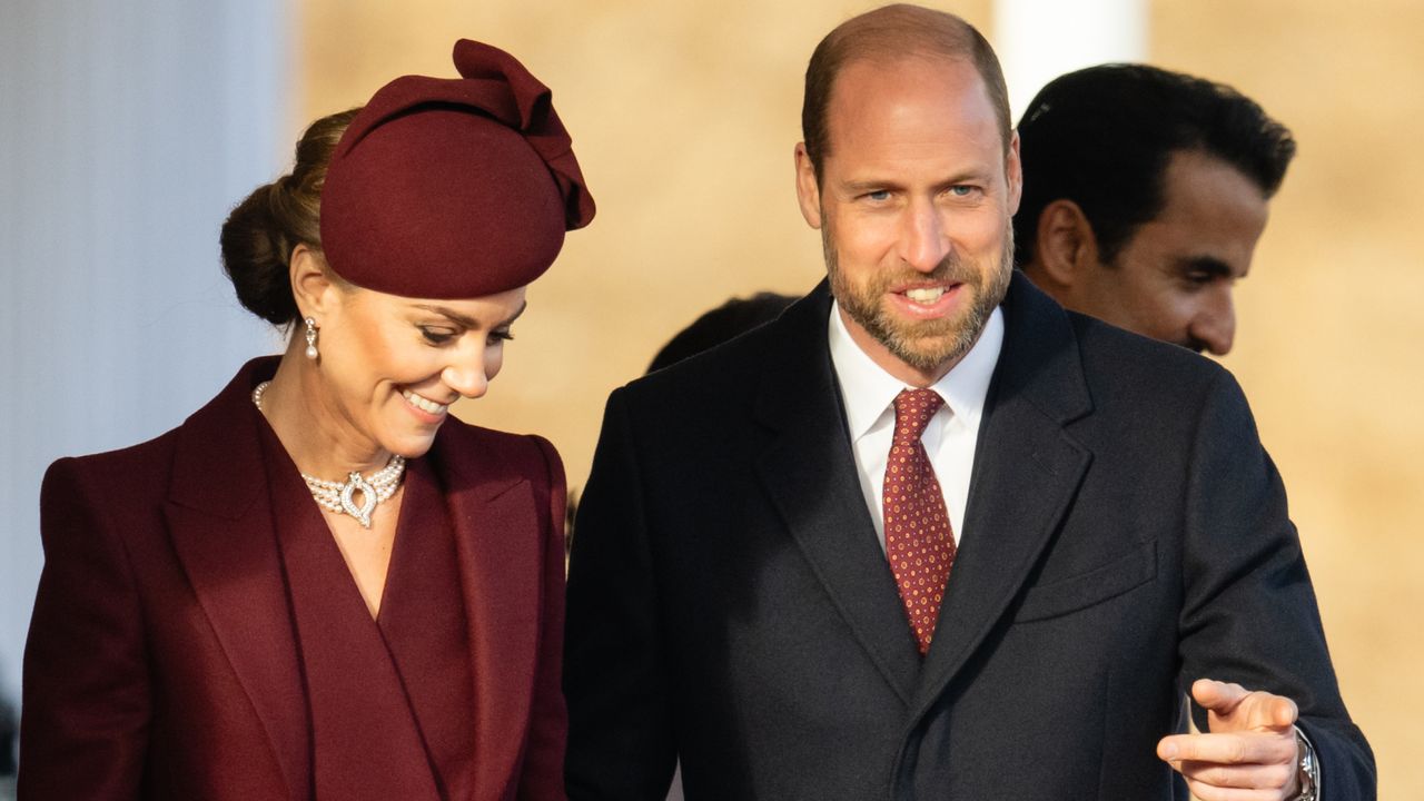 Princess Kate wearing a maroon coat and hat standing next to Prince William and smiling 