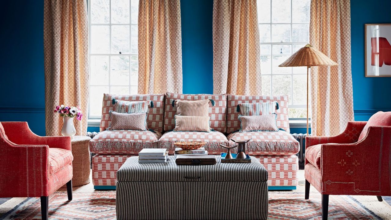 Blue painted living room with patterned curtains, pink sof and chairs, striped ottoman, rattan floor lamp, patterned rug, single artwork on wall