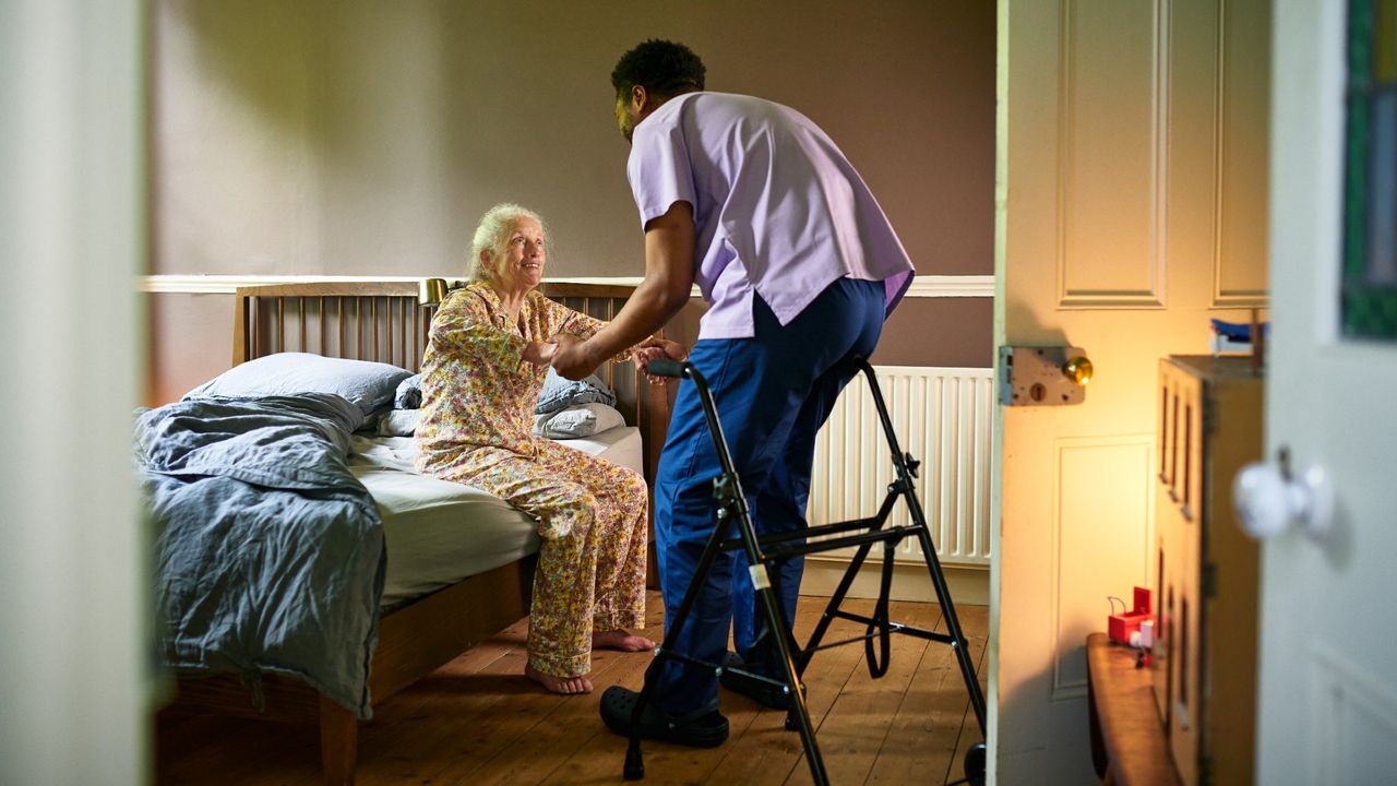 Young male care worker helping elderly woman off bed in a nursing home